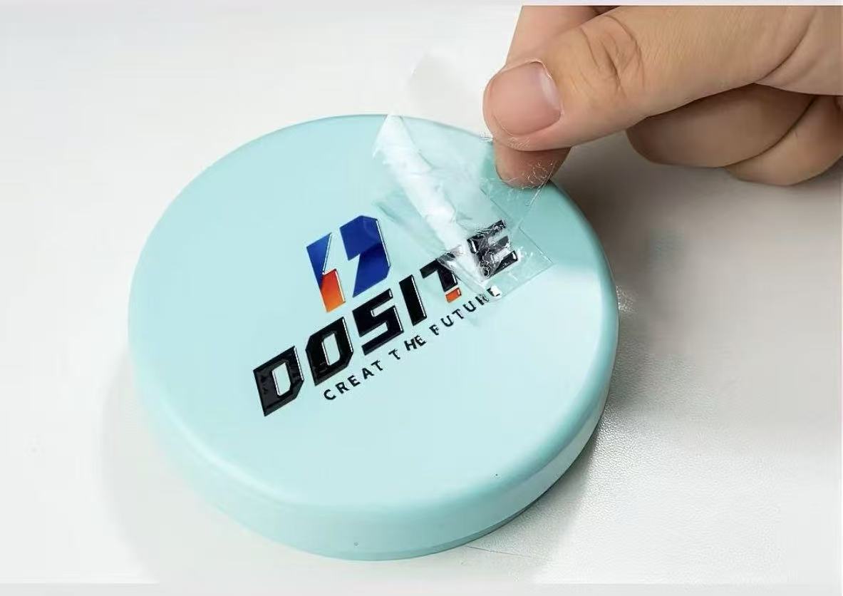Woven label with a black and red logo 'DOSITE CREATE THE FUTURE' on a circular light blue product, being peeled off.